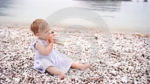 Little girl drinks water from a bottle while sitting on a pebble beach by the sea