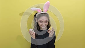 little girl in Easter bunny ears holding two painted easter eggs on a yellow