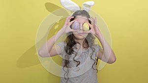 little girl in Easter bunny ears holding two painted easter eggs on a yellow