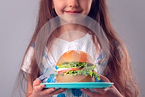 Little girl eating big burger. Kid looking at healthy big sandwich, studio isolated on white background
