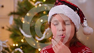 Little girl eating Christmas cookies near the Christmas tree. Happy Child in a Santa hat and a red sweater