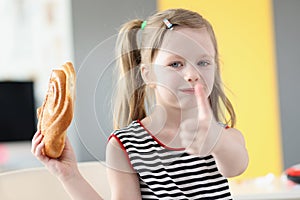 Little girl eating delicious sweet bun and showing thumb up