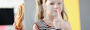 Little girl eating delicious sweet bun and showing thumb up