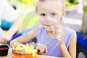 Little girl eating rench fries with sauce at street cafe outside.
