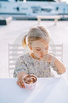 Little girl eats ice cream with a spoon from a cup while sitting at the table