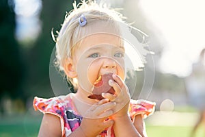 Little girl eats a peach on a green lawn holding it by hands. Portrait