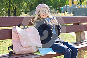 Little girl elementary school student sitting on bench with backpack, looks at school books and notebooks. Background autumn city