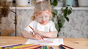 A little girl enthusiastically draws a heart with colored pencils sitting at the table at home alone