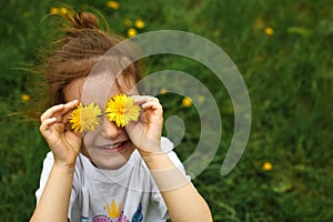 A little girl of European appearance with light hair puts yellow dandelion flowers to her eyes and enjoys the summer,