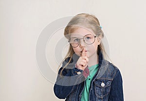 Little girl in eyeglasses putting finger up to lips and ask silence. Child  doing a Please Keep Quiet gesture towards the camera