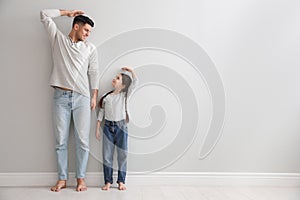 Little girl and father measuring their height near light grey wall indoors. Space for text