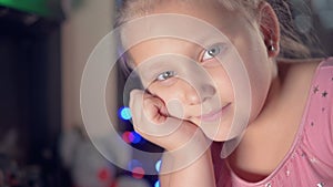 A little girl in a festive dress against the backdrop of festive lights and a Christmas tree smily looks at camera blur