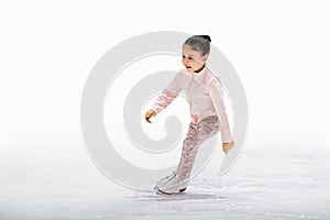 Little girl figure skater in light pink tracksuit with smile skates on the ice on an indoor skating rink
