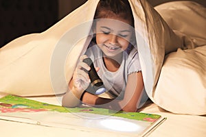 Little girl with flashlight reading book under blanket