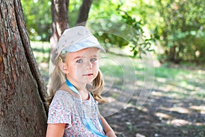 Little girl in a floral dress and hat leaning against a tree in the park