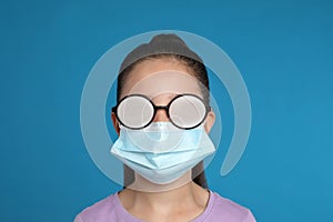 Little girl with foggy glasses caused by wearing medical face mask on blue background. Protective measure during coronavirus