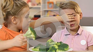 Little girl fooling brother, giving to eat broccoli instead of sweets, pranking
