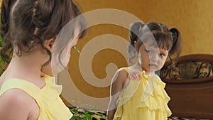 The little girl in front of the mirror bites her lip. A beautiful child in a yellow dress by the mirror. Girl with tails.