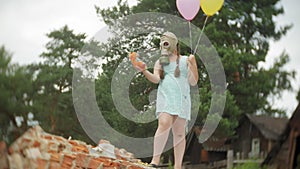 A little girl in a gas mask on the ruins of a building and holding on to a doll and balloons.