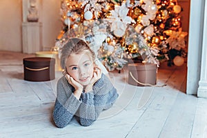 Little girl with gift box near Christmas tree on Christmas eve at home. Young kid in light bedroom with winter