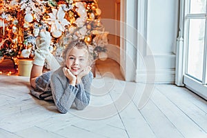 Little girl with gift box near Christmas tree on Christmas eve at home. Young kid in light bedroom with winter