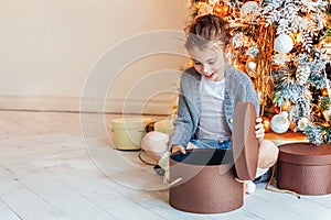 Little girl with gift box near Christmas tree on Christmas eve at home. Young kid in light bedroom with winter decoration.