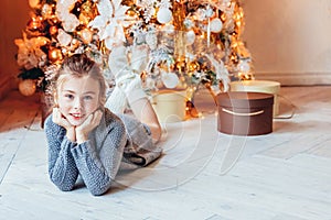 Little girl with gift box near Christmas tree on Christmas eve at home. Young kid in light bedroom with winter decoration Happy