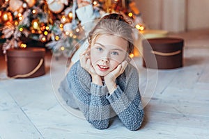 Little girl with gift box near Christmas tree on Christmas eve at home. Young kid in light bedroom with winter decoration Happy