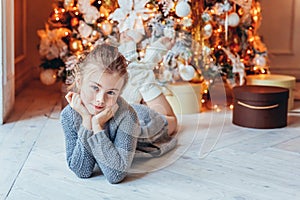 Little girl with gift box near Christmas tree on Christmas eve at home. Young kid in light bedroom with winter decoration Happy