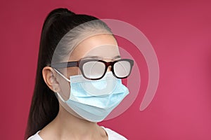 Little girl with glasses caused by wearing medical face mask on pink background. Protective measure during coronavirus