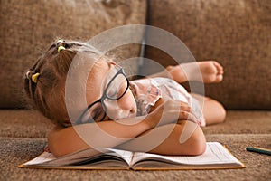 A little girl with glasses fell asleep on the couch reading a book while studying at home