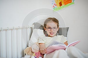 Little girl in glasses reading a book while lying in bed.