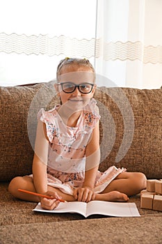 A little girl with glasses sits on the couch and studies. Studying at home