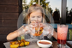 a little girl with gloves eats a burger and potatoes in a summer outdoor cafe.