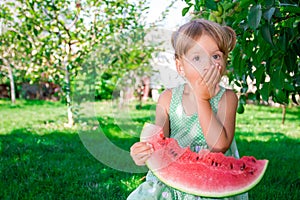 Little girl in green dress, barefoot standing in the park with big slice watermelon.