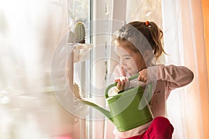 A little girl with a green watering can is playing on the window of the house and watering the flowers.