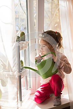 A little girl with a green watering can is playing on the window of the house and watering the flowers.