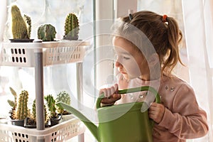 A little girl with a green watering can is playing on the window of the house and watering the flowers.