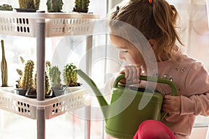 A little girl with a green watering can is playing on the window of the house and watering the flowers.