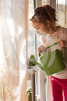 A little girl with a green watering can is playing on the window of the house and watering the flowers.