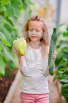 Little girl in greenhouse with pepper and cucumber in hands. Time to harvest. Big basket full of vegetables