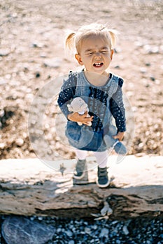 Little girl grimaces while standing on a snag with toys on the beach