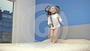 Little girl with a hairbrush in her hands jumps on a large parental bed. Child has a lot of fun.