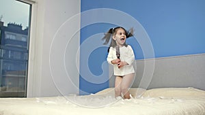 Little girl with a hairbrush in her hands jumps on a large parental bed. Child has a lot of fun.