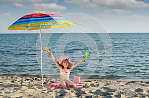 Little girl with hands up sitting under sunshade on beach