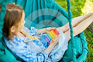 A little girl the on hanging chair outdoors play pop it, kid hands playing with colorful pop It
