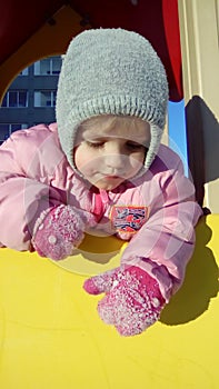 A little girl hangs down a wooden slide and looks down. The child is wearing a gray hat, a pink thick jacket, raspberry mittens