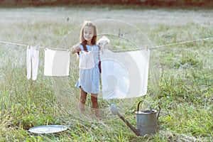 Little girl hangs out to dry retro doll clothes, summer nature outdoor. Washing, children`s games, kid`s leisure