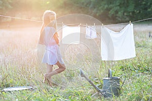 Little girl hangs out to dry retro doll clothes, summer nature outdoor. Washing, children`s games, kid`s leisure