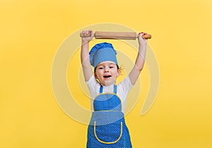 a little girl is happy in a kitchen apron and a hat with a rolling pin on a yellow background. Looking at the camera